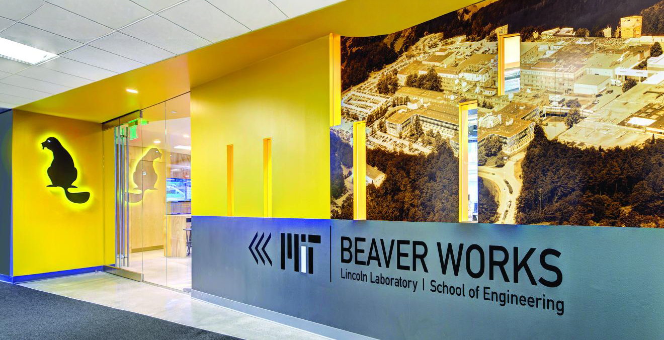 Entrance to Beaver Works' facility. The image shows a yellow wall with a glass door. There is a backlit beaver logo to the left of the door and on the right is an aerial photo in sepia colors along with a logo lockup with the words MIT, Beaver Works, Lincoln Laboratory and School of Engineering.