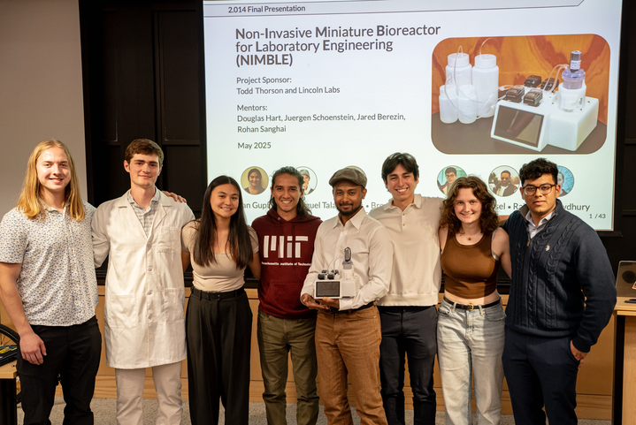 A group of eight people standing together in front of a presentation slide titled “Non-Invasive Miniature Bioreactor for Laboratory Engineering (NIMBLE).” They are smiling and posing with a small scientific device, while the slide behind them shows project details and images of laboratory equipment.