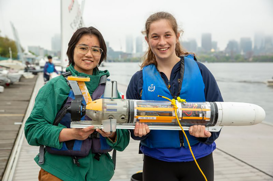 Two students wearing life jackets stand on a dock holding a small underwater vehicle on the Charles River in Boston, with the city skyline in the background.