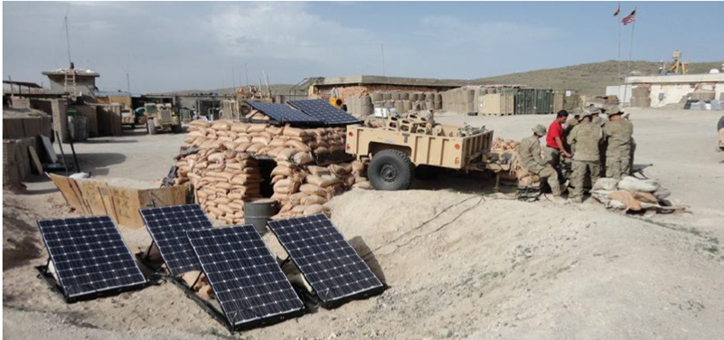 Several solar panels are set up on the ground and on top of a sandbag-covered shelter at a military outpost. A trailer is positioned nearby, and a group of soldiers is gathered to the right side of the image. American flags are visible in the background.