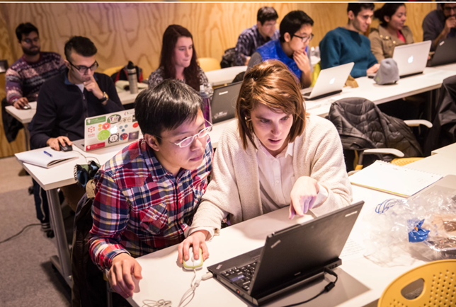 Two students looking at a laptop. Behind them are other students in a classroom setting.