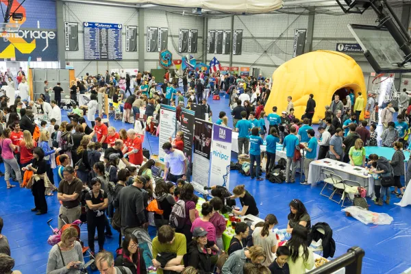 Photo shows a wide view of a gymnasium during the Cambridge Science Festival, with many tables set up for experiments. Each table is manned by several volunteers. Hundreds of people enjoying the hands-on experiments at each booth.