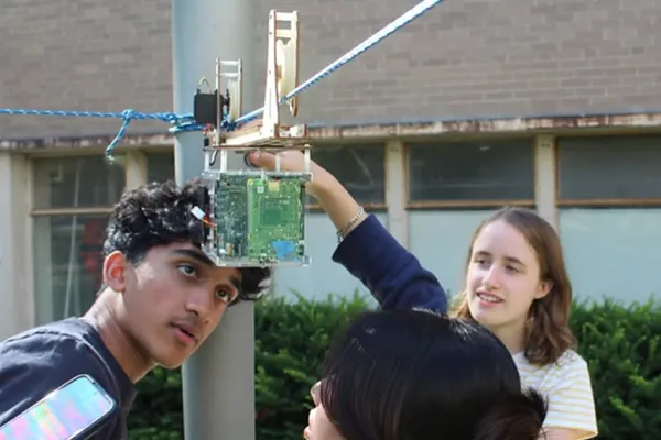 Three students inspect their self-built CubeSat (a plastic cube containing a computer board programmed to take photos) as it hangs from a rope and pulley system to mimic an orbital path while it takes pictures of “Earth” below.