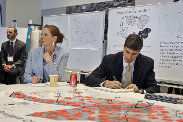 A female executive is poised to take notes as she listens during a workshop on Humanitarian Assistance and Disaster Relief held at MIT Beaver Works. A male executive takes notes. The table is filled with reports and a map with movable squares indicating planning involved for each location. In the background is a whiteboard with a mission statement, a process design flow, and a map highlighting specific regions needing aid.