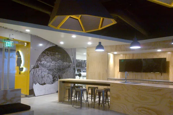 The photo shows a cafe area with bar stools and tv screens. A yellow wall on the left side features the Beaver works logo lit up from behind.