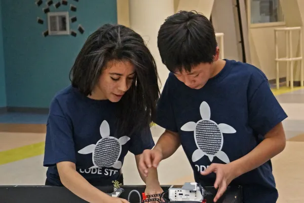 A 6th grade girl and boy in matching robotics team t-shirts are adjusting components on a shoebox-sized robotic car made of LEGO bricks and controlled by their self-coded programming.