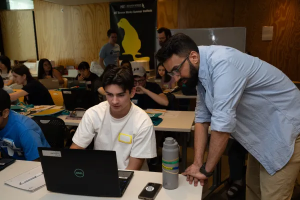 In a small classroom, a student is working on his laptop computer. A mentor leans over at the edge of the table, helping the student go through the proper steps to build a secure encryption scheme online.