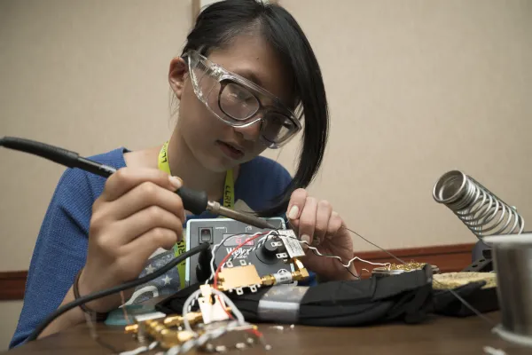 A 12th grade girl wearing safety glasses solders wires on a circuit board in place.