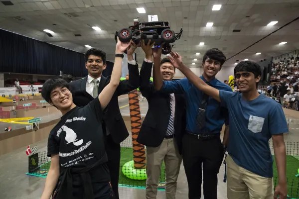 A team of five students jubilantly raise their self-built, self-programmed autonomous mini-car after successfully completing the racetrack obstacle course shown behind them. 