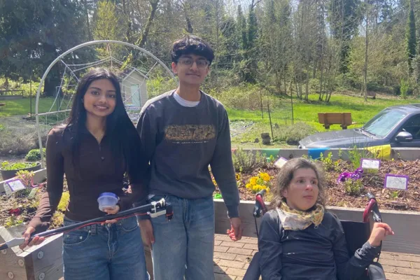 A high school boy and girl stand in a garden with their client, who has reduced grip capacity and limited range of motion. The team is showing the adaptive tool they built to help their client take care of plants in her garden. 