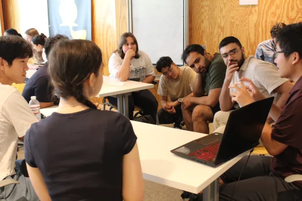 A group of seven high-school students in the Beaver Works Summer Institute program listen intently to a college-aged teacher's assistant explains an engineering concept. The teacher's assistant has his laptop open and facing the students to help them understand how the concept is applied to creating their project. 