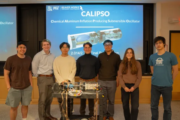 A team of 7 students is shown on stage after making a presentation about their creation: a buoyant engine that uses reversible hydrogen fuel cell to adjust buoyancy. The engine (named CALIPSO, for Chemical Aluminum Inflation Producing Submersible Oscillator)  is shown on stage with the introductory slide of their presentation shown in the background.