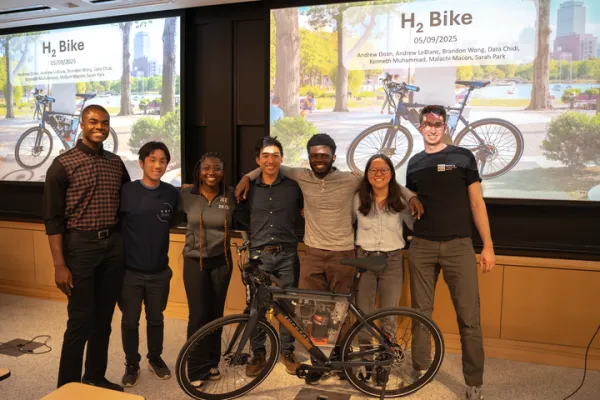 A group of seven people standing together in front of a presentation slide titled “H₂ Bike” with images of a bicycle by a waterfront. They are posing with a black bicycle equipped with technical components on display in front of them.