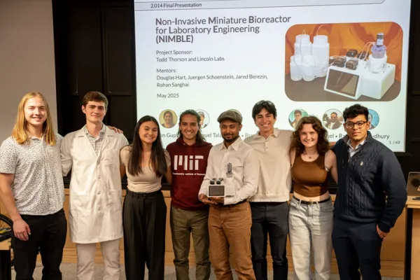 A group of eight people standing together in front of a presentation slide titled “Non-Invasive Miniature Bioreactor for Laboratory Engineering (NIMBLE).” They are smiling and posing with a small scientific device, while the slide behind them shows project details and images of laboratory equipment.