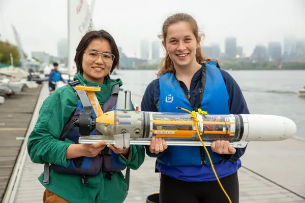 Two students wearing life jackets stand on a dock holding a small underwater vehicle on the Charles River in Boston, with the city skyline in the background.