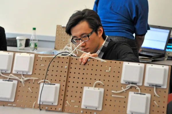 A student sits at a work table adjusting wires on two particle boards screwed together. Each board holds student-built flat box-shaped radar systems that work together and can be pointed at objects to evaluate thickness, erosion, structural flaws, and weather patterns.