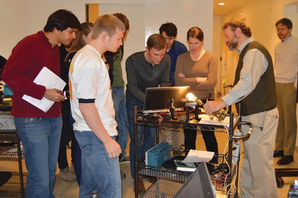As group of seven college students test their self-built laser communication terminal, an instructor shows how a bright light source (in this case, a lightbulb) interferes with the signal the terminal is trying to detect. 
