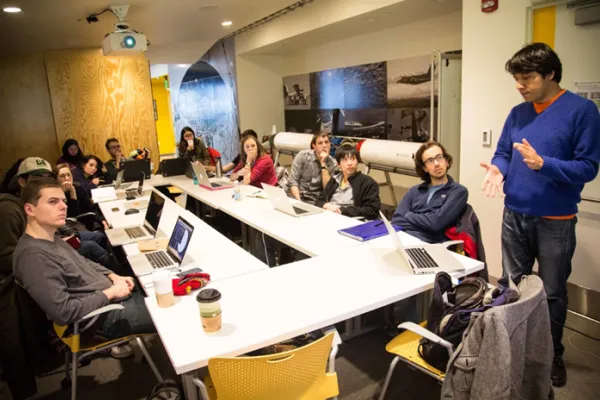 A 14-person class sits at a long table in a Beaver Works classroom. Each student has their laptop in front of them and looks to a projected screen as the instructor gives a presentation. 
