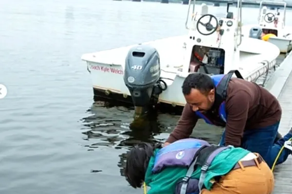 Two students hold a self-built autonomous underwater vehicle that looks like a mini submarine in a plastic casing. The students help the prototype descend into the Charles River for field tests. 