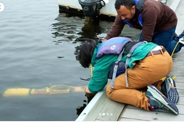 Two students hold a self-built autonomous underwater vehicle that looks like a mini submarine in a plastic casing. The students help the prototype descend into the Charles River for field tests.